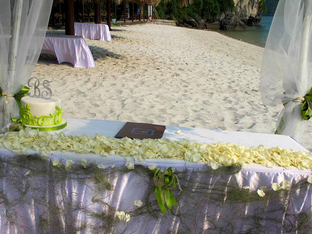 Bàn cử hành lễ được trang trí bởi những cánh hoa hồng trắng. Chiếc bánh cưới trắng với hoa văn xanh và chữ cái tên cô dâu chú rể cũng được đặt riêng.